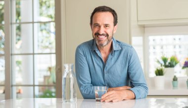 Orta yaş adam ayakta ve diş gösterilen kendine güvenen bir gülümsemeyle gülümseyerek mutlu bir yüz ile bir bardak su içme