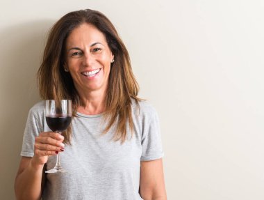 Orta yaş kadın ayakta ve diş gösterilen kendine güvenen bir gülümsemeyle gülümseyerek mutlu bir yüz ile bir bardak kırmızı şarap içme