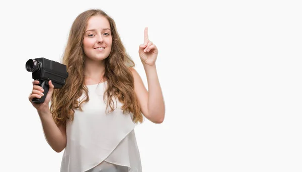 スーパー カメラを保持している若いブロンドの女性が幸せな顔で ナンバーワンのアイデアや質問ポインティング指で驚いた — ストック写真