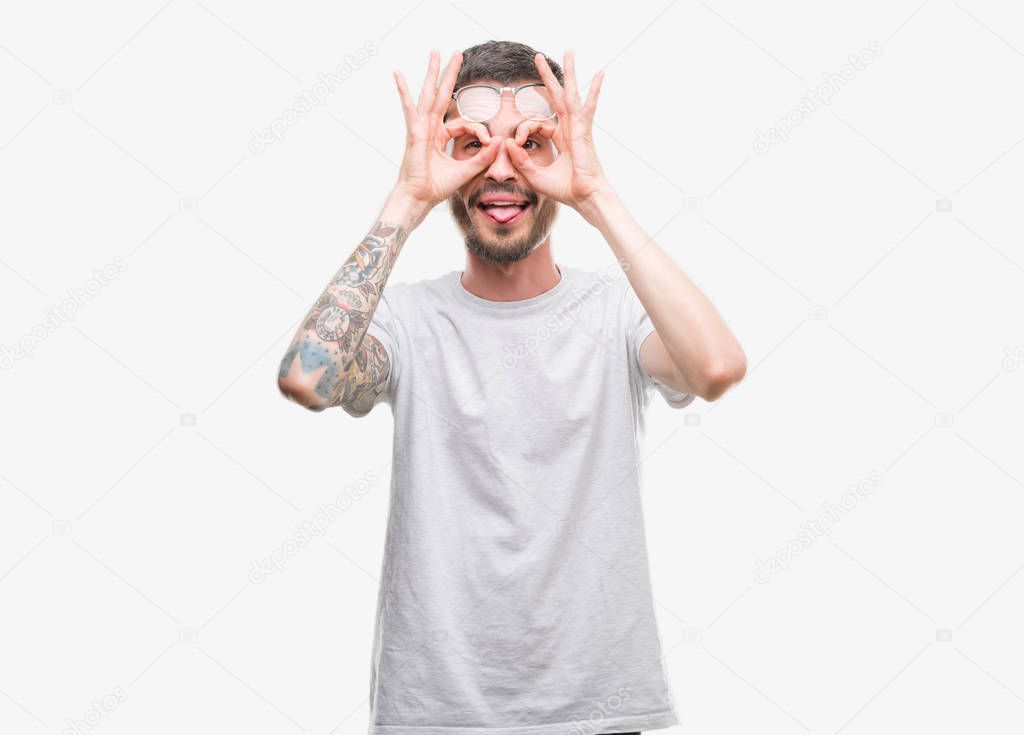 Young tattooed adult man doing ok gesture like binoculars sticking tongue out, eyes looking through fingers. Crazy expression.