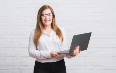 Genç Yetişkin iş kadını ayakta ve diş gösterilen kendine güvenen bir gülümsemeyle gülümseyerek mutlu bir yüz ile bilgisayar dizüstü holding beyaz tuğla duvarın üzerinden