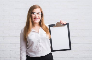 Ayakta ve diş gösterilen kendine güvenen bir gülümsemeyle gülümseyerek mutlu bir yüz ile beyaz tuğla duvar holding Pano üzerinde duran genç yetişkin iş kadın