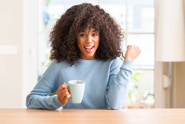 African american woman holding a cup of coffee at home screaming proud and celebrating victory and success very excited, cheering emotion