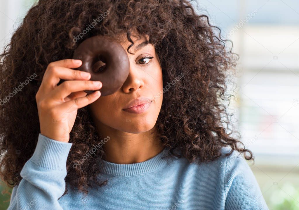 Young african american woman holding chocolate donut at home with a confident expression on smart face thinking serious