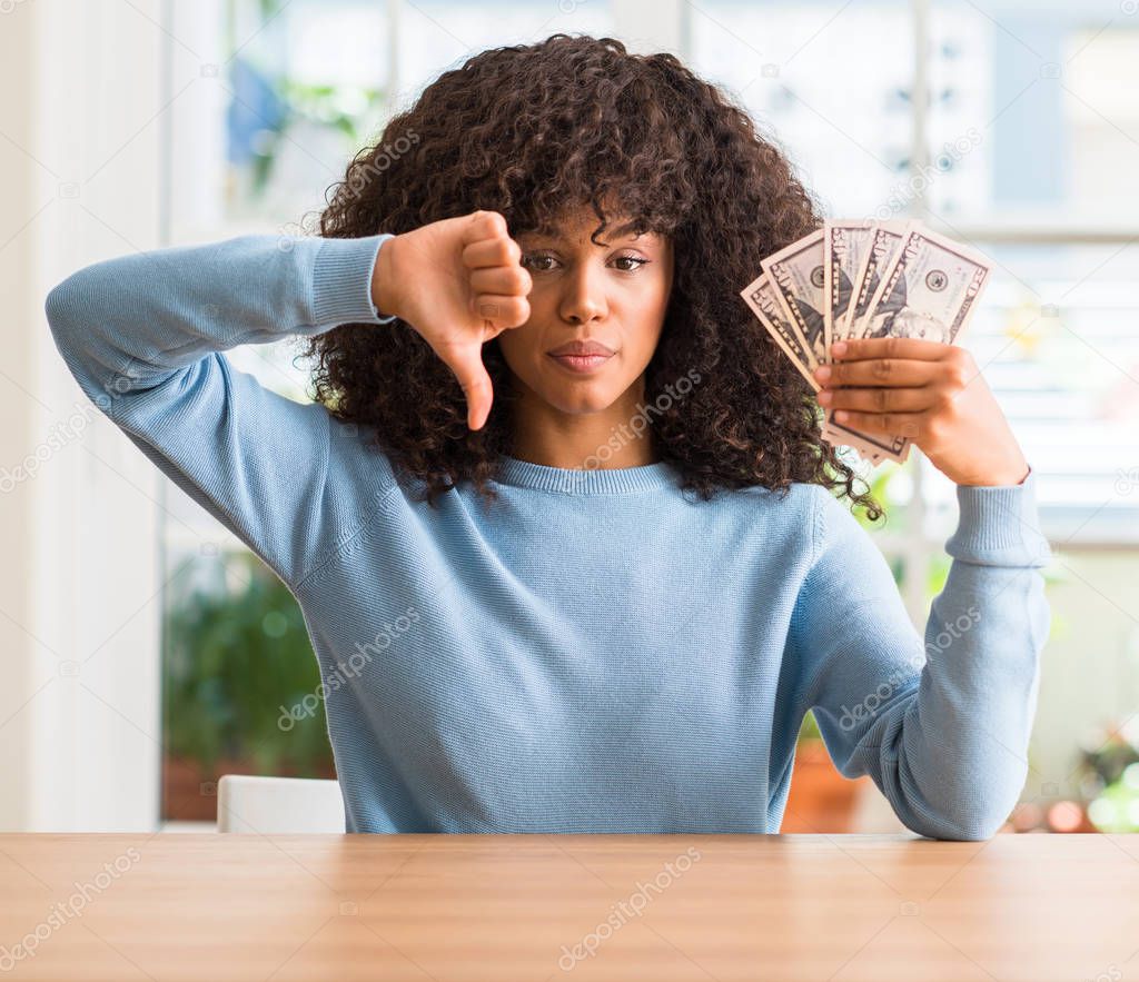 African american woman holding dollar bank notes with angry face, negative sign showing dislike with thumbs down, rejection concept
