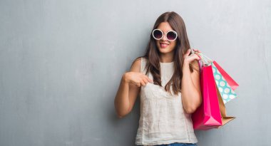 Grunge gri duvar sürpriz yüz işaret parmağı renkli alışveriş torbaları için kendini tutan genç esmer kadın