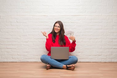 Holding dolar el ve parmak ile tarafına işaret çok mutlu ödeme beyaz tuğla duvarın üzerinden katta oturan genç esmer kadın