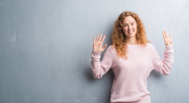 Gri grunge genç Kızıl saçlı kadın duvar gösterilen giymiş pembe kazak ve parmakları ile işaret numara 9 kendinden emin ve mutlu gülümseyerek süre.