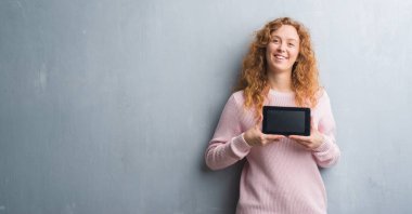 Ayakta ve diş gösterilen kendine güvenen bir gülümsemeyle gülümseyerek mutlu bir yüz ile tablet kullanarak gri grunge duvar genç Kızıl saçlı kadın