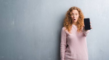 Gri grunge duvar gösteren smartphone ekran şok sürpriz yüzü, korkuyor ve korku ifadesi ile heyecanlı olan korkmuş genç Kızıl saçlı kadın