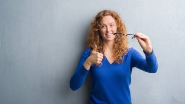 Genç Kızıl saçlı kadın optik gözlük ile mutlu büyük holding gri grunge duvarın üzerinden gülümseme ok işareti, başparmak ile parmak, mükemmel işareti yapıyor