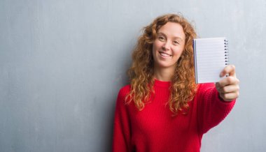 Gri grunge duvar gösteren boş defter ayakta ve diş gösterilen kendine güvenen bir gülümsemeyle gülümseyerek mutlu bir yüz ile genç Kızıl saçlı kadın