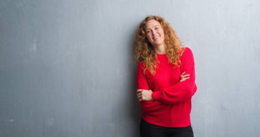 Kameraya bakarak çapraz silah ile gülümseyen genç redhead gri grunge duvar giyen üzerinde kırmızı kadın kazak gülen surat. Olumlu bir kişi.
