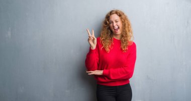 Mutlu yüz zafer işareti yaparken kameraya göz kırpıyor ile gülümseyen genç redhead gri grunge duvar giyen üzerinde kırmızı kadın kazak. İki numaralı.