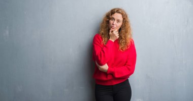 Genç redhead gri grunge duvar giyen üzerinde kırmızı kadın kazak emin çapraz kollar ve çeneme kaldırdı el ile gülümseme ile kameraya bakıyor. Pozitif düşünce.