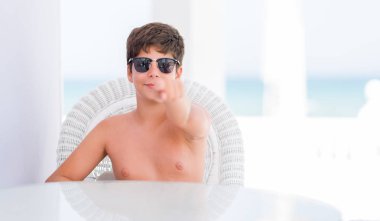 Genç çocuk kamera ve size parmak ile işaret sahilde bir sandalyede oturan tatil günlerinde el işareti, pozitif ve kendinden emin bir jest ön