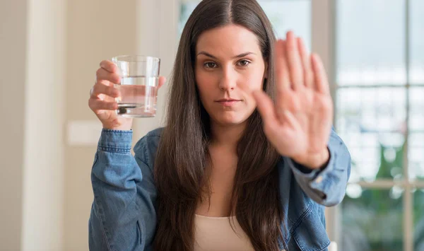 年轻妇女喝玻璃水在家用张开手做停止标志以严肃和自信的表示 防御手势 — 图库照片