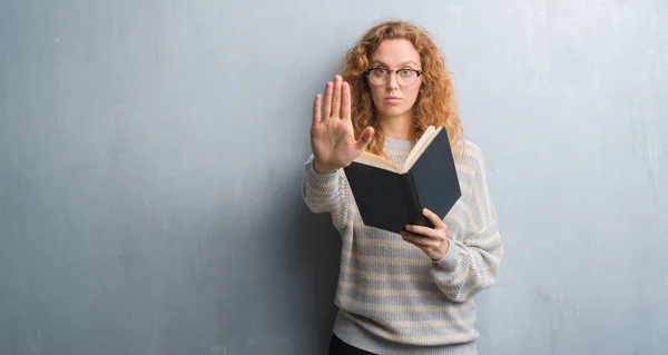 灰色垃圾墙上的年轻红头发的女人读一本书用张开手做停止标志以严肃和自信的表达 防御手势 — 图库照片