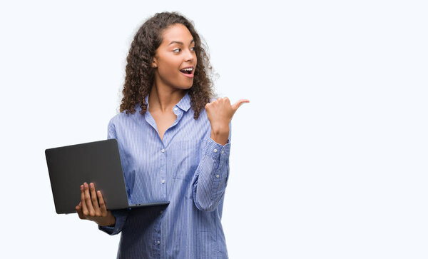 Young hispanic woman holding computer laptop pointing and showing with thumb up to the side with happy face smiling