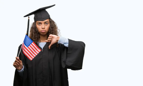 Young hispanic woman wearing graduation uniform holding flag of United States with angry face, negative sign showing dislike with thumbs down, rejection concept