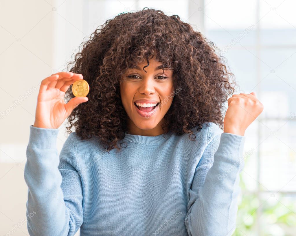 African american woman holding golden bitcoin cryptocurrency at home screaming proud and celebrating victory and success very excited, cheering emotion