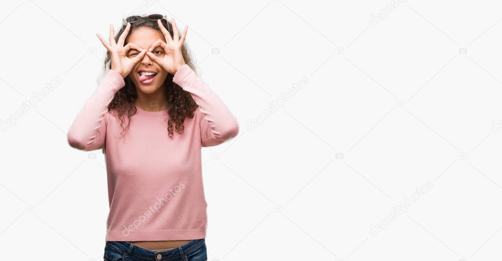 Beautiful young hispanic woman wearing sunglasses doing ok gesture like binoculars sticking tongue out, eyes looking through fingers. Crazy expression.