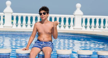 Genç çocuk tatilde plajda işaret ve başparmak kadar mutlu yüz gülümseyen tarafı ile gösterilen yüzme havuzunda