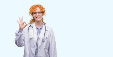 Doktor üniforması el ve parmaklar ok işaretiyle yapıyor olumlu gülümseyen genç Kızıl saçlı kadın. Başarılı ifade.