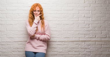 Emin çapraz kollar ve çeneme kaldırdı el ile gülümseme ile kameraya bakarak tuğla duvarın üzerinden duran genç Kızıl saçlı kadın. Pozitif düşünce.