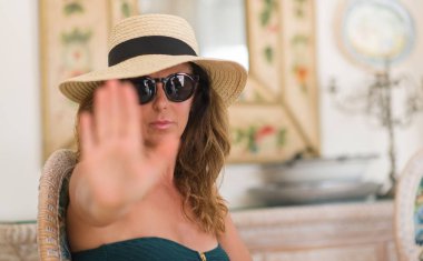 Orta yaş esmer kadın giyiyor yaz şapka tatil ile açık el dur işareti ile ciddi ve kendine güvenen ifade, savunma hareketi yapıyor