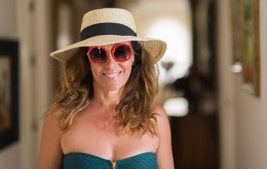 Orta yaş esmer kadın ayakta ve diş gösterilen kendine güvenen bir gülümsemeyle gülümseyerek mutlu bir yüz ile güneş gözlüğü takıyor