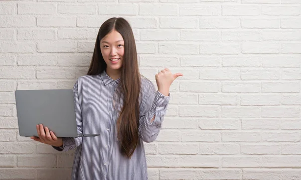 年轻的中国妇女在砖墙使用膝上型电脑指向和显示与拇指到一边 快乐的脸微笑着 — 图库照片