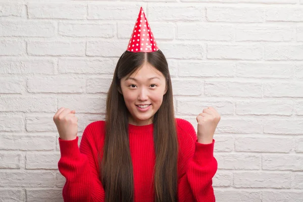 感情を応援して誇りに思って叫んで 勝利と非常に興奮して 成功を祝う誕生日の帽子を身に着けているレンガの壁の上の若い中国の女性 — ストック写真