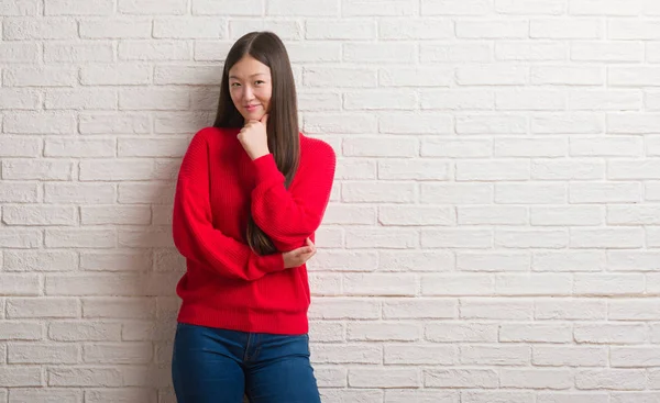 Young Chinese woman over brick wall looking confident at the camera with smile with crossed arms and hand raised on chin. Thinking positive.