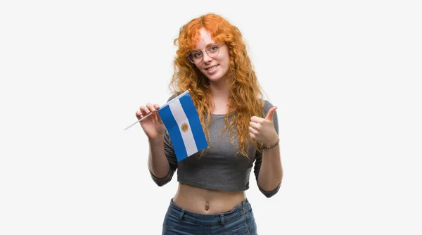優秀な兆候の指で サインをしている笑顔に満足してアルゼンチンの若い赤毛の女性持株旗が親指 — ストック写真