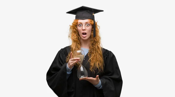 年轻红发学生女子身穿毕业制服抱沙漏吓坏了惊讶的脸 害怕和激动与恐惧的表情 — 图库照片