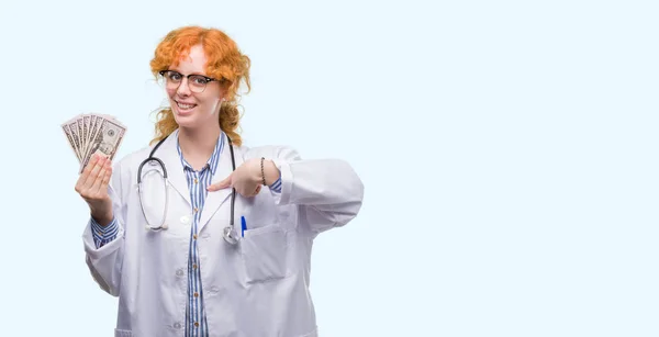 Young Redhead Doctor Woman Holding Dollars Surprise Face Pointing Finger — Stock Photo, Image