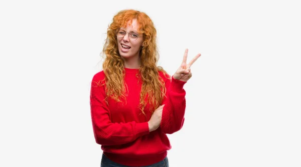 勝利のサインを行うカメラでウィンクしている幸せそうな顔を浮かべて赤いセーターを着ている若い赤毛の女性 — ストック写真