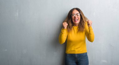 Orta yaş İspanyol kadın kolları olan başarı için heyecanlı gözlüklü gri duvar yüzünden zafer kutluyor gülümseyen kaldırdı. Kazanan kavramı.