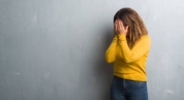 Orta yaş İspanyol kadın ağlayarak süre elleriyle yüzünü kapsayan üzgün ifade ile gözlüklü gri duvar yüzünden. Depresyon kavramı.