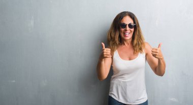 Orta yaş İspanyol kadın güneş gözlüğü başarı işareti el ile olumlu hareketi yaparken giyiyor gri duvar yüzünden başparmak gülümseyen ve mutlu. Neşeli bir ifadeyle, kazanan jest kameraya bakıyor.