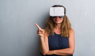 Sanal gerçeklik gözlük el ve parmak ile tarafına işaret çok mutlu gri grunge duvarın üzerinden duran orta yaş İspanyol kadın