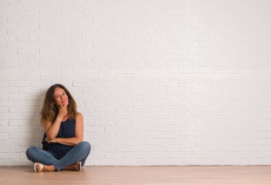 Orta yaş İspanyol kadın emin çapraz kollar ve çeneme kaldırdı el ile gülümseme ile kameraya bakarak beyaz tuğla duvarın üzerinden yere oturma. Pozitif düşünce.