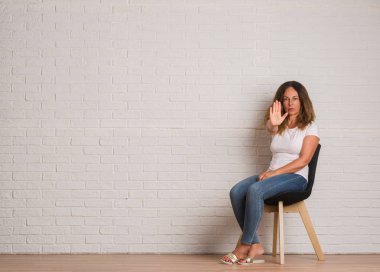 Orta yaş İspanyol kadın iskemlede beyaz tuğla walll ile üzerinde dur işareti ile ciddi ve kendine güvenen ifade, savunma hareketi yaparken el açmak