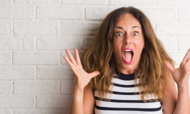 Orta yaş İspanyol kadın beyaz tuğla duvar çok mutlu ve heyecanlı, kocaman bir gülümseme ile çığlık ve ellerini kaldırdı kazanan ifade kutluyor zafer