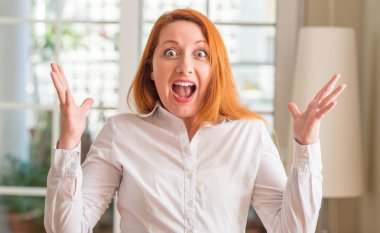 Evde çok mutlu ve heyecanlı, beyaz gömlek giyen kızıl saçlı kadın büyük gülümseme ile çığlık ve ellerini kaldırdı kazanan ifade kutluyor zafer