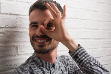 Genç yetişkin adam mutlu yüz el parmaklarının arasından seyir göz üzerinde ok işaretiyle yapıyor gülümseyen ile beyaz tuğla duvarı ayakta