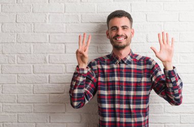 Genç yetişkin adam gösterilen ve parmakları ile işaret beyaz tuğla duvarın üzerinden ayakta sekiz kendinden emin ve mutlu gülümseyerek iken numara.