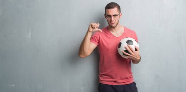 Futbol futbol topuyla kızgın yüzü, eksi işareti aşağı antipati başparmak ile gösterilen, ret kavramı holding gri grunge duvarın üzerinden beyaz delikanlı