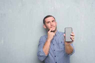 Gri grunge duvar gösteren boş ekran smartphone ciddi bir yüz düşünme hakkında soru, çok karışık fikir genç beyaz adam